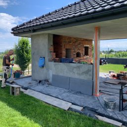 Pan - Budowa zewnętrznego kominka z cegły: murarz zamiata teren. Widoczne narzędzia, cegły, elementy konstrukcji oraz otoczenie ogrodu. Prace w toku, dzień.