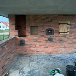 Pan - Murowany piec zewnętrzny z cegły klinkierowej pod zadaszeniem, z elementami żeliwnymi i drewnianymi drzwiczkami. Widoczne narzędzia i materiały budowlane.