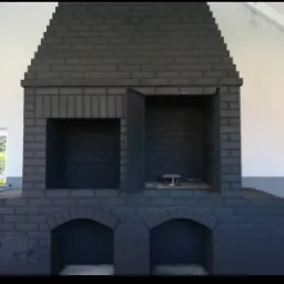 Szary, murowany kominek zewnętrzny z otwartymi drzwiczkami, widoczny z przodu na tle białej ściany. Widoczne elementy konstrukcyjne i detale wykończenia.