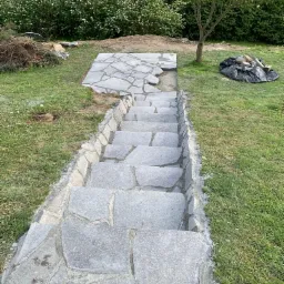 Kamienne schody ogrodowe w trakcie budowy, z widocznymi elementami krajobrazu i narzędziami. Częściowo ułożona nawierzchnia z nieregularnych płyt.