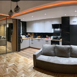 Pan - Panoramiczne ujęcie wnętrza po remoncie: nowoczesna kuchnia z czarno-białymi płytkami, parkiet w jodełkę, lustrzane drzwi przesuwne i sofa na pierwszym planie.