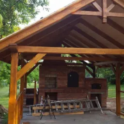 Zadaszona konstrukcja z drewnianymi belkami, pod którą znajduje się murowany z cegły piec z dwoma otworami, drabina oparta o ścianę, w tle zieleń.