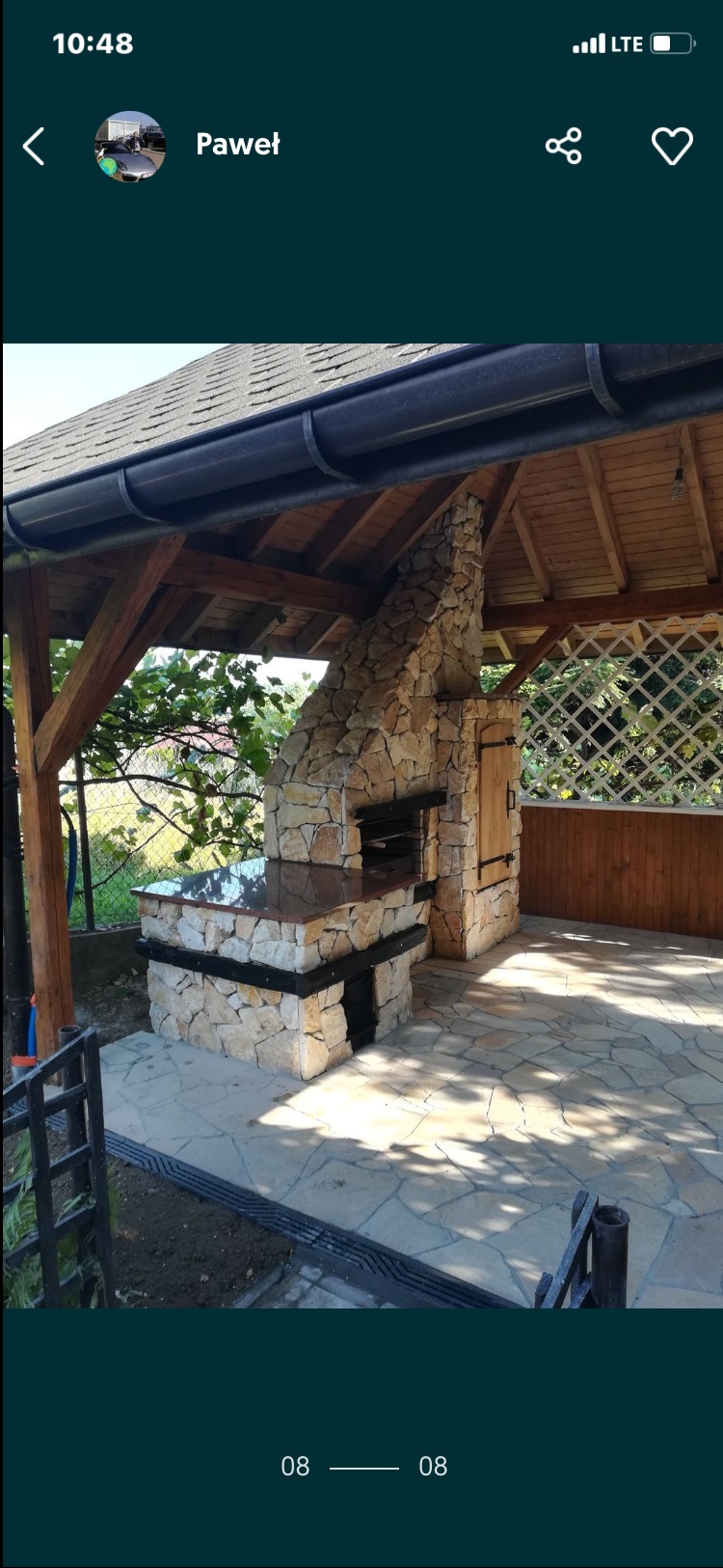 Kamienny kominek zewnętrzny pod zadaszeniem z drewnianą konstrukcją i kamienną posadzką. Widoczne elementy grilla i wędzarni z drewnianymi drzwiczkami.