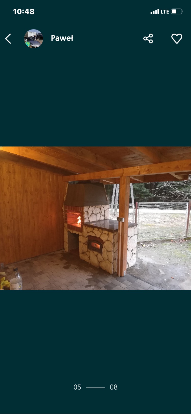 Zadaszona, kamienna kuchnia letnia z paleniskiem i piecem chlebowym. Widoczne płomienie w palenisku, drewniana konstrukcja dachu i ściany. Tło stanowi ogrodzenie i zieleń.