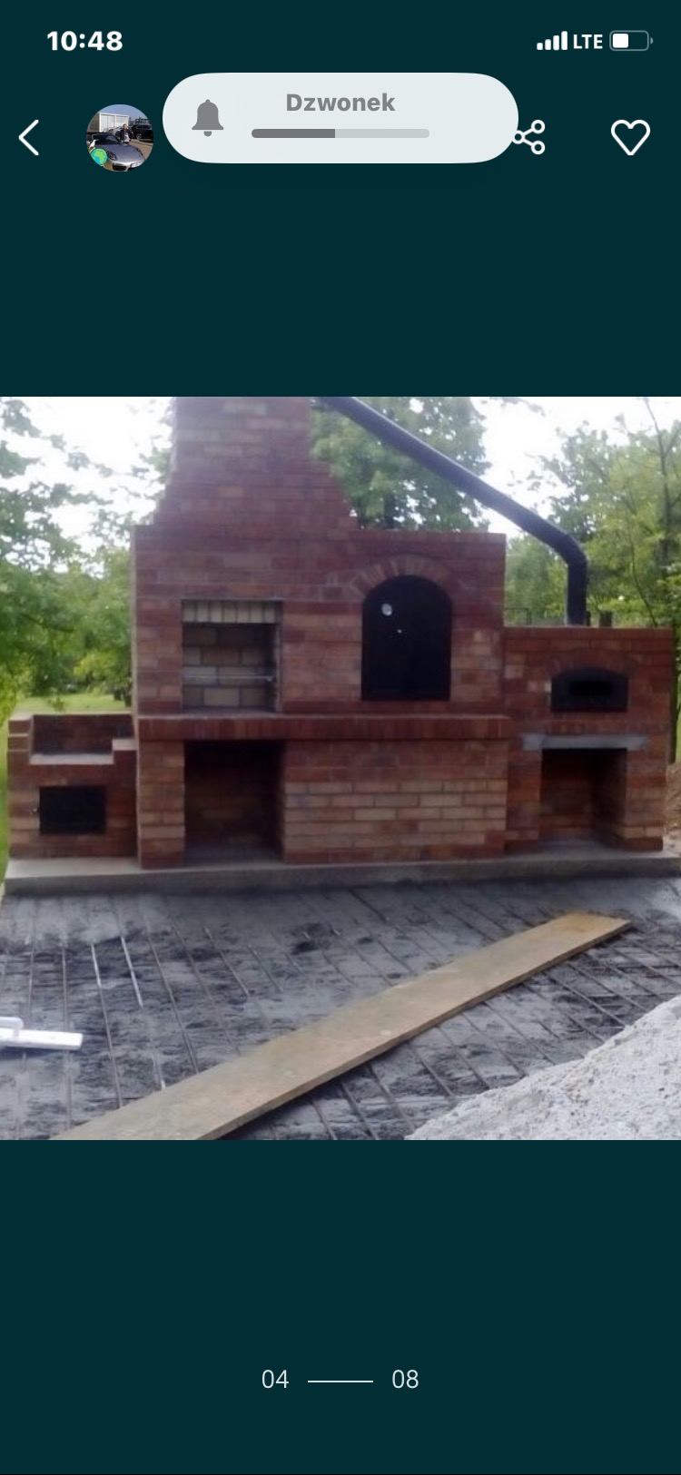 Murowany kominek zewnętrzny z cegły, z czarnym kominem, z widocznym zbrojeniem podłoża. Elementy rusztu i deska na pierwszym planie.