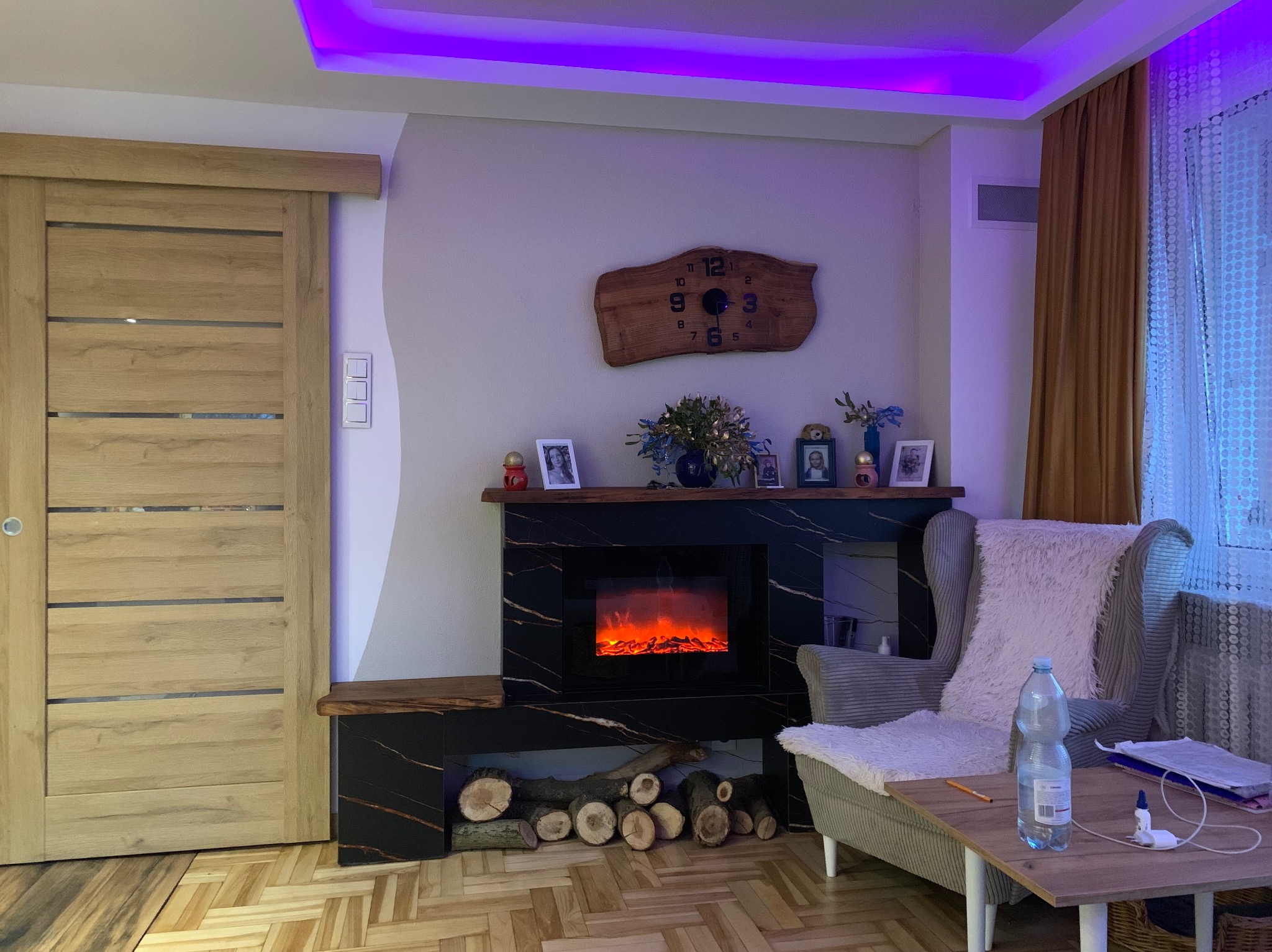 Salon z kominkiem elektrycznym obudowanym czarnym marmurem, drewniane drzwi przesuwne, drewniany zegar na ścianie, fotel z narzutą i stolik kawowy.