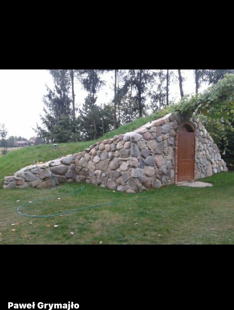 Ziemianka z kamienia polnego, częściowo pokryta darnią, z drewnianymi drzwiami. Otoczenie: trawnik, drzewa iglaste i liściaste. Nietypowa konstrukcja.