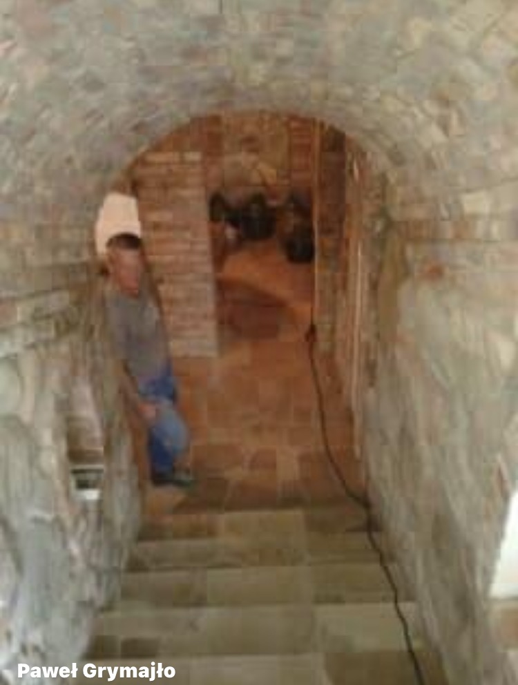 Murowany łuk z cegły, schody z kamienia, mężczyzna w kasku. Kamienna konstrukcja przypomina piwnicę lub podziemne przejście. Widoczne fugi i naturalna kolorystyka materiałów.