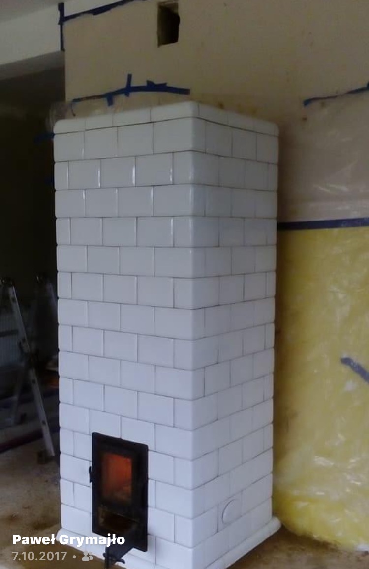 Biały piec kaflowy w trakcie budowy, widoczne zabezpieczenia ścian folią i taśmą malarską, niedokończone wykończenie sufitu.
