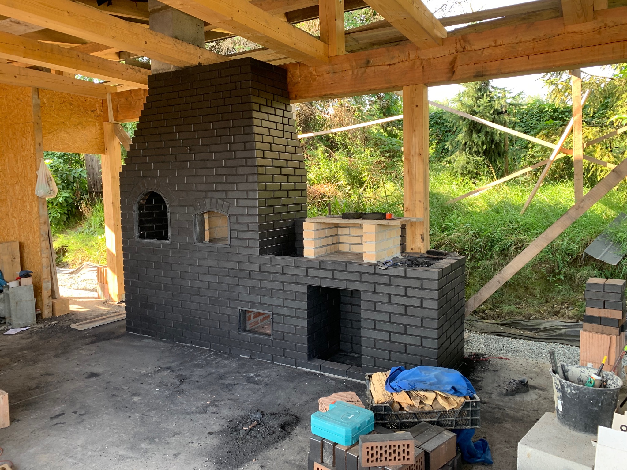 Ciemna, ceglana konstrukcja kominka zewnętrznego w trakcie budowy, z widocznymi elementami szkieletu drewnianego i narzędziami murarskimi wokół. Detal ceglanego wykończenia.