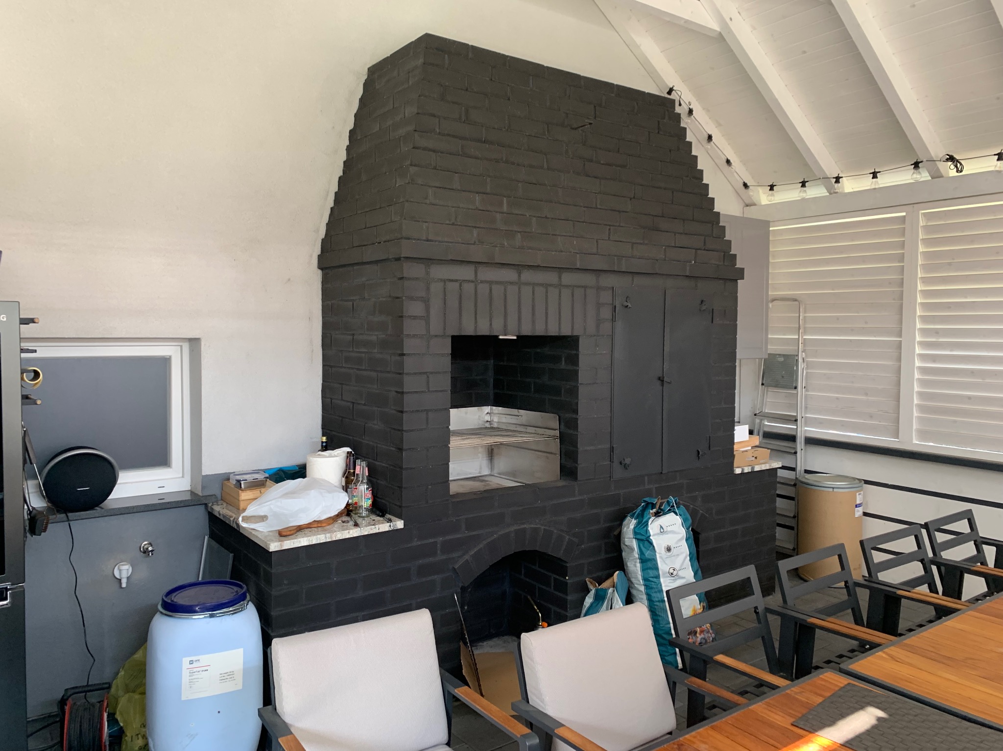 Murowany, czarny kominek zewnętrzny z grillem w zadaszonej przestrzeni. Widoczne krzesła i stół. Oświetlenie w stylu girlandy.