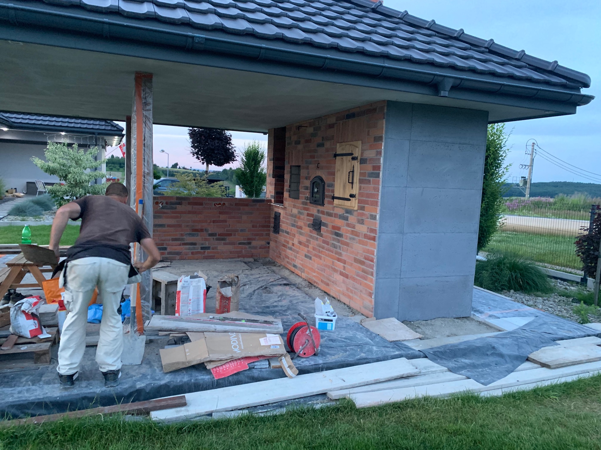 Budowa pieca chlebowego z cegły na zewnątrz. Widoczny murarz w trakcie pracy, narzędzia i materiały budowlane. Częściowo zadaszona konstrukcja z ciemną dachówką.