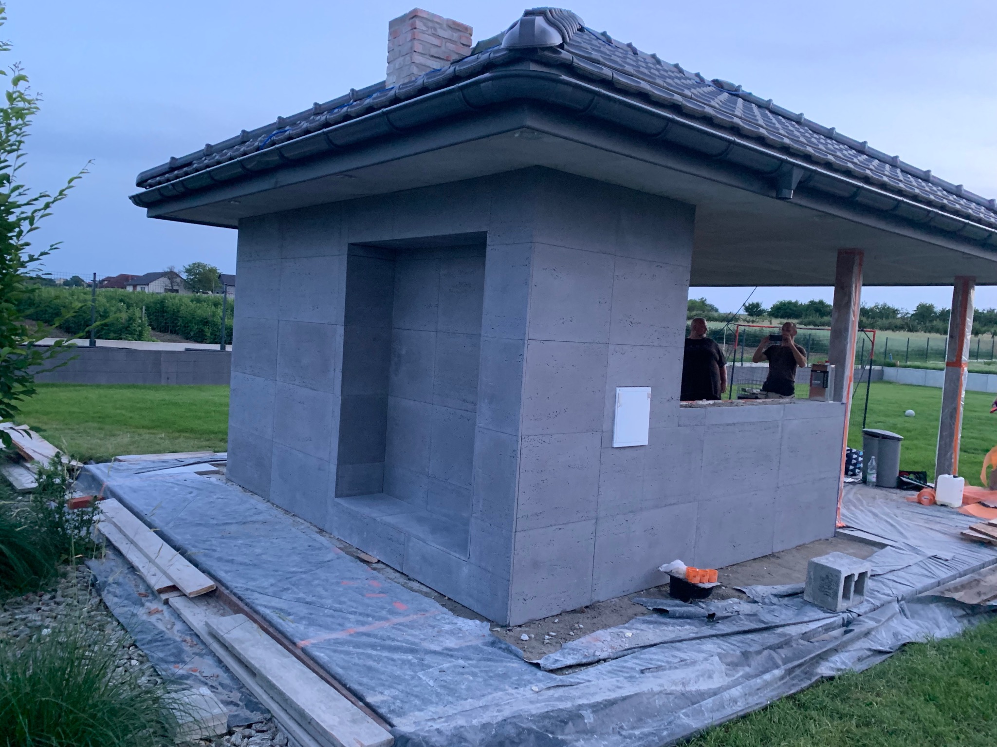 Betonowa altana z kominem w trakcie budowy, widoczne płyty elewacyjne, drewniane elementy konstrukcyjne i narzędzia. Dwóch mężczyzn w tle.