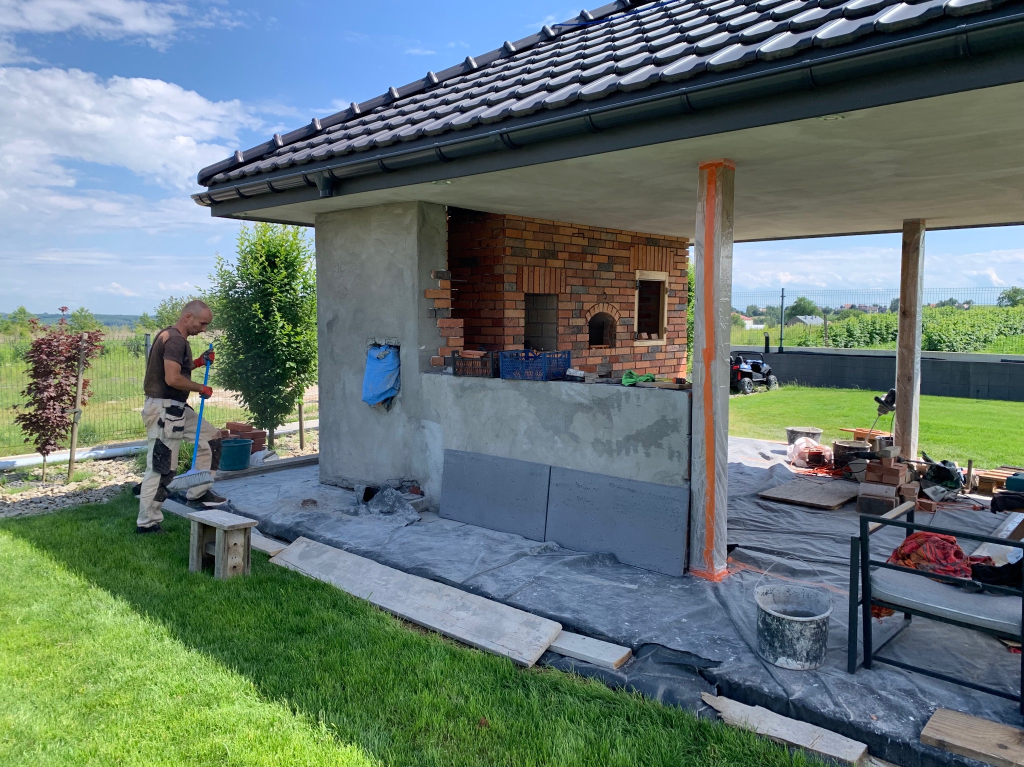 Budowa zewnętrznego kominka z cegły: murarz zamiata teren. Widoczne narzędzia, cegły, elementy konstrukcji oraz otoczenie ogrodu. Prace w toku, dzień.