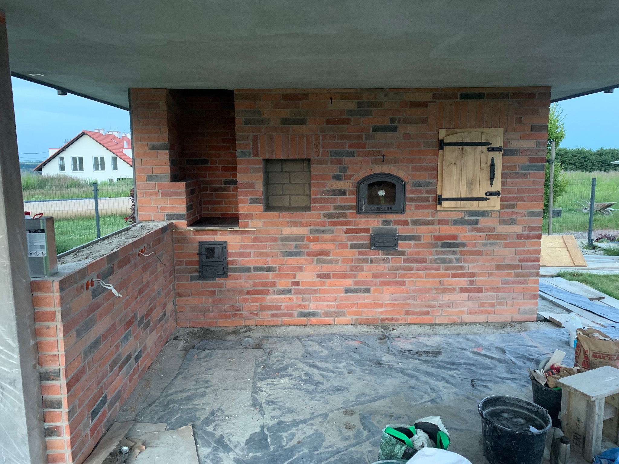 Murowany piec zewnętrzny z cegły klinkierowej pod zadaszeniem, z elementami żeliwnymi i drewnianymi drzwiczkami. Widoczne narzędzia i materiały budowlane.