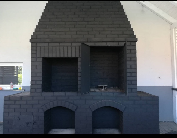 Szary, murowany kominek zewnętrzny z otwartymi drzwiczkami, widoczny z przodu na tle białej ściany. Widoczne elementy konstrukcyjne i detale wykończenia.