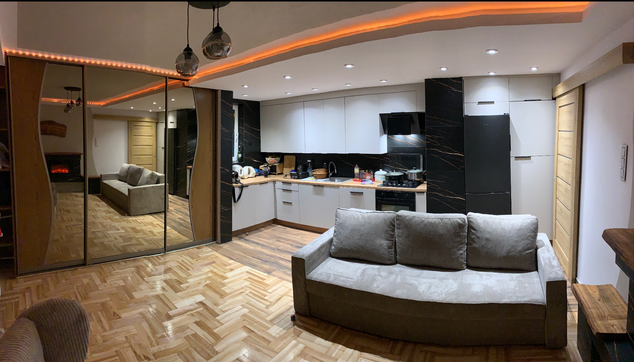 Panoramiczne ujęcie wnętrza po remoncie: nowoczesna kuchnia z czarno-białymi płytkami, parkiet w jodełkę, lustrzane drzwi przesuwne i sofa na pierwszym planie.