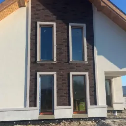 Nowoczesny dom z elewacją w kolorze złamanej bieli i fragmentem z cegły klinkierowej, z czterema oknami z białymi obramowaniami. W tle góry i błękitne niebo.