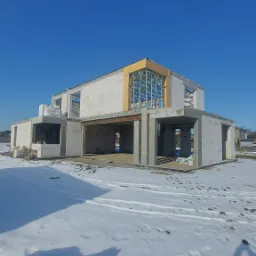 Niedokończony dwukondygnacyjny budynek z betonu komórkowego w zimowej scenerii, z widoczną konstrukcją stalową w miejscu okna na górnej kondygnacji i śniegiem na pierwszym planie.