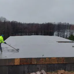 Wyrównywanie wylewki fundamentowej na budowie domu jednorodzinnego, widok na las i jezioro w tle, pracownik w odblaskowej kurtce używa łaty wibracyjnej.