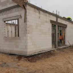 Wznoszenie ścian parterowego domu z białych bloczków, widoczne okna i przygotowane otwory drzwiowe, na górze ściany wylany wieniec, na zewnątrz widoczne ślady prac ziemnych.