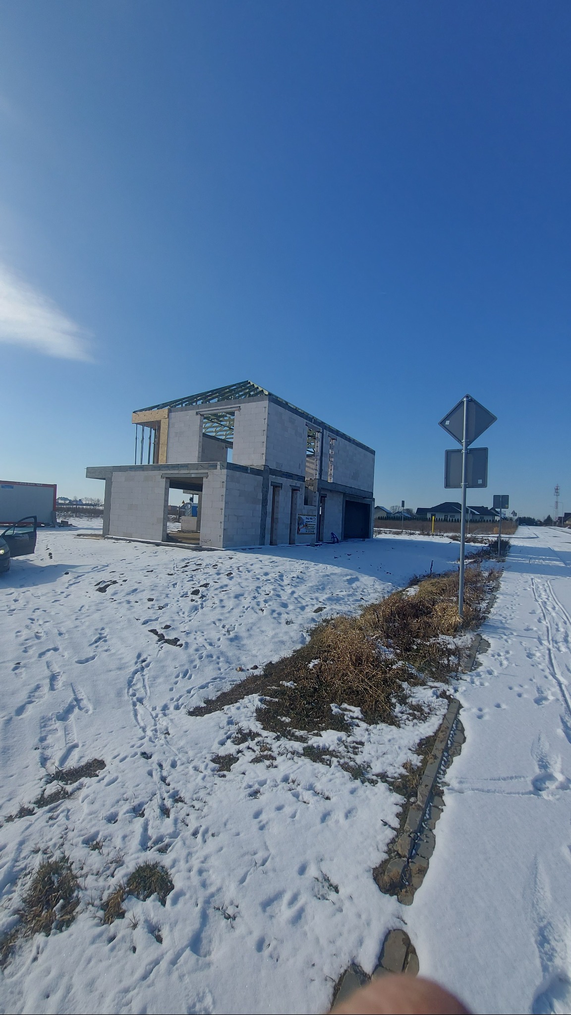 Surowy stan dwukondygnacyjnego budynku z bloczków, z widoczną konstrukcją dachu, na zasypanym śniegiem terenie w słoneczny dzień. Widoczny znak drogowy i fragment samochodu w tle.