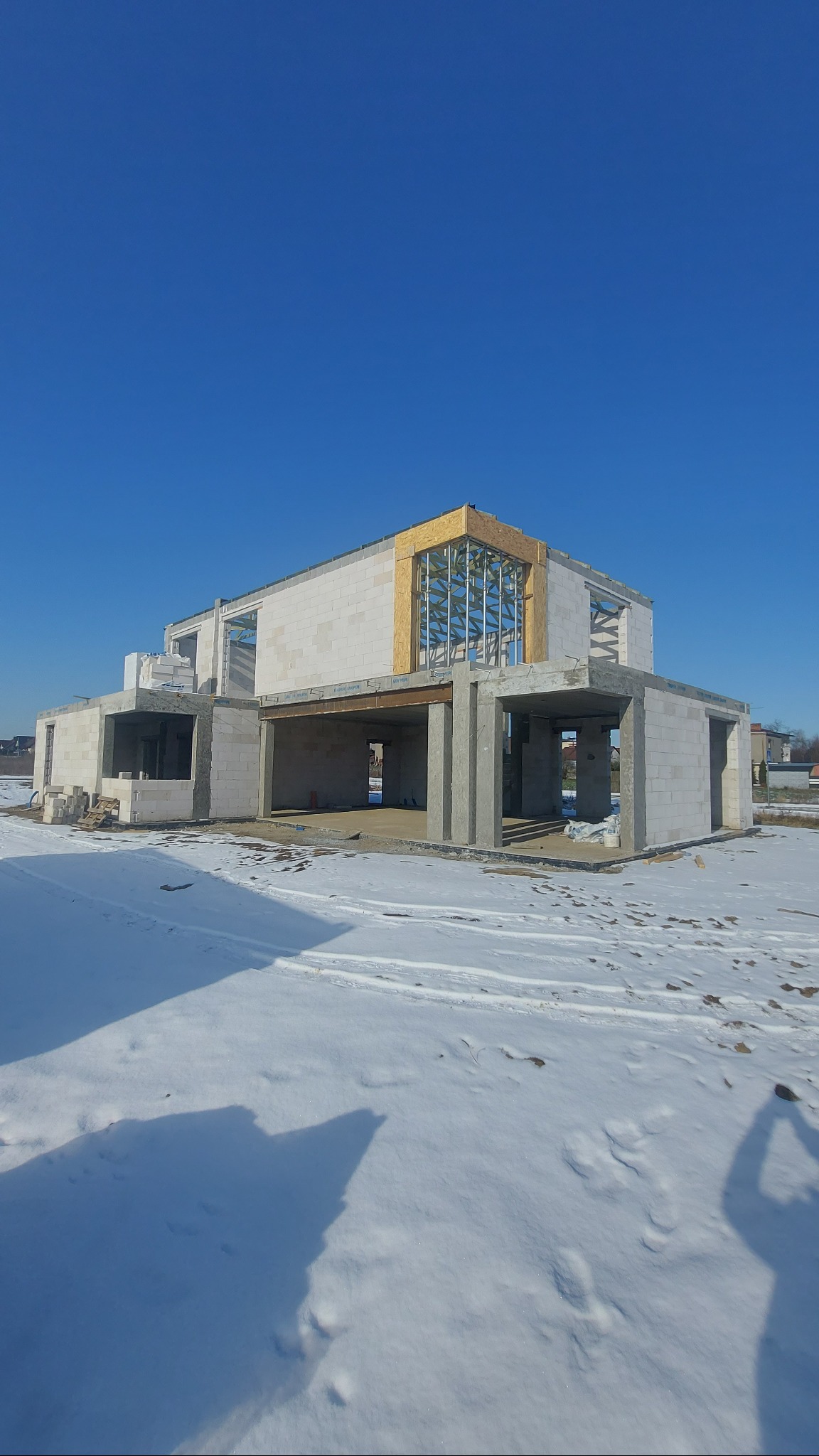 Niedokończony dwukondygnacyjny budynek z betonu komórkowego w zimowej scenerii, z widoczną konstrukcją stalową w miejscu okna na górnej kondygnacji i śniegiem na pierwszym planie.