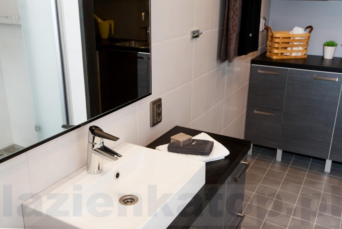 Bateria umywalkowa Oras Optima 2710F na białej umywalce, obok mydło i ręcznik na czarnym blacie w łazience z szarą podłogą i szafkami.