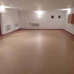 Beżowa wykładzina PCV z bordową obwódką w przestronnym, jasnym pomieszczeniu z białymi ścianami, grzejnikami i drewnianymi drzwiami.