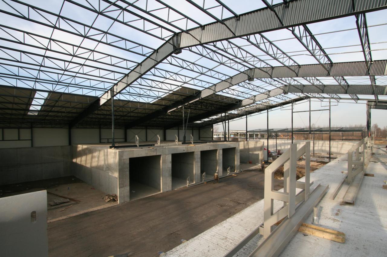 Wnętrze hali w trakcie budowy z widoczną stalową konstrukcją dachu i betonowymi elementami. Surowy stan wykończenia, widoczne detale konstrukcyjne.
