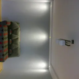 Nowoczesny salon z szarą sofą w geometryczne wzory, oświetleniem punktowym w suficie i minimalistyczną lampą ścienną. Ujęcie z g&oacute;ry, podkreślające detale wykończenia.