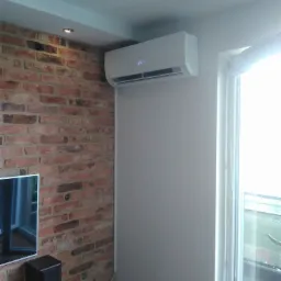 Biały klimatyzator ścienny z wyświetlaną temperaturą 24 stopnie, zamontowany w rogu pokoju z ceglaną ścianą i oknem balkonowym.