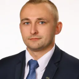 Portret mężczyzny w garniturze z przypinką OVB, na białym tle. Profesjonalny wygląd, niebieski krawat i jasne oczy.