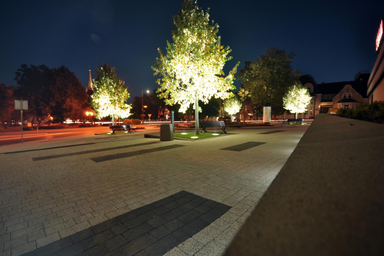 Nocny widok na oświetlony plac miejski z drzewami, ławkami i brukowaną nawierzchnią. Perspektywa z dołu, ciemne niebo w tle.