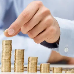 Dłoń mężczyzny w błękitnej koszuli układa złotą monetę na szczycie stosu monet, tworząc rosnący wykres.