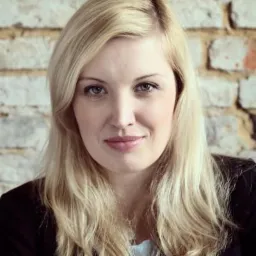 Portret uśmiechniętej blondynki w czarnej marynarce na tle ściany z jasnej cegły.