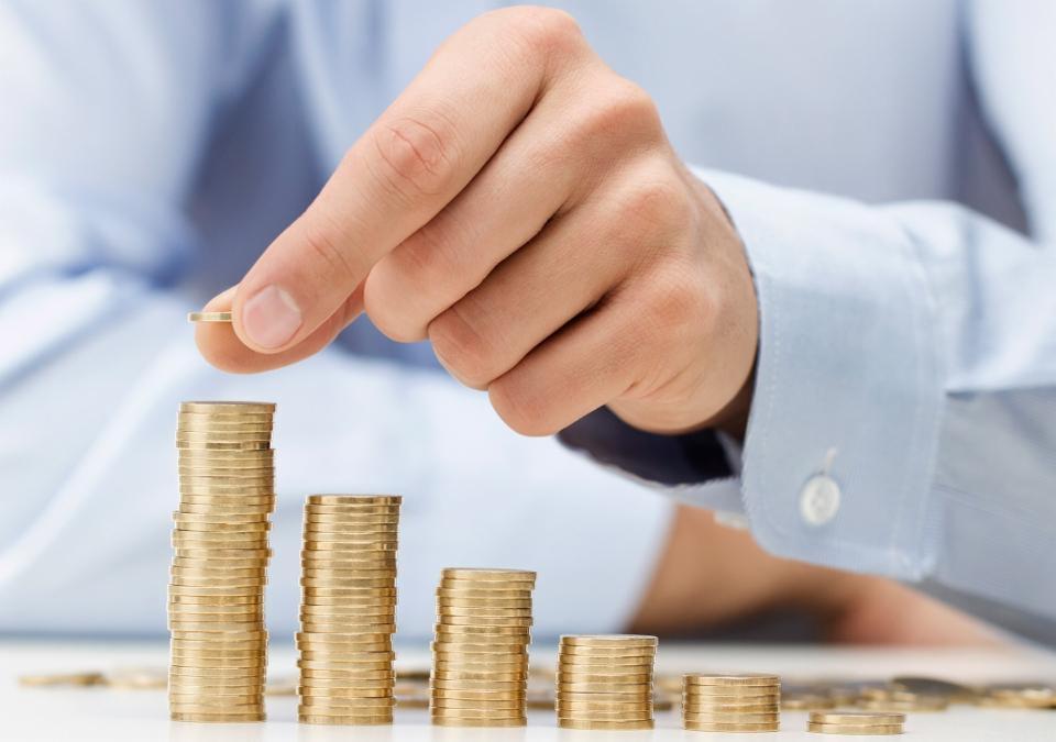 Dłoń mężczyzny w błękitnej koszuli układa złotą monetę na szczycie stosu monet, tworząc rosnący wykres.
