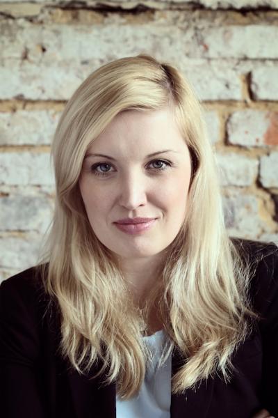 Portret uśmiechniętej blondynki w czarnej marynarce na tle ściany z jasnej cegły.