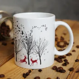 Biały kubek z nadrukiem zimowego krajobrazu: sylwetki drzew, stado ptaków i dwa czerwone jelenie, otoczony rozsypanymi ziarnami kawy na drewnianym stole.