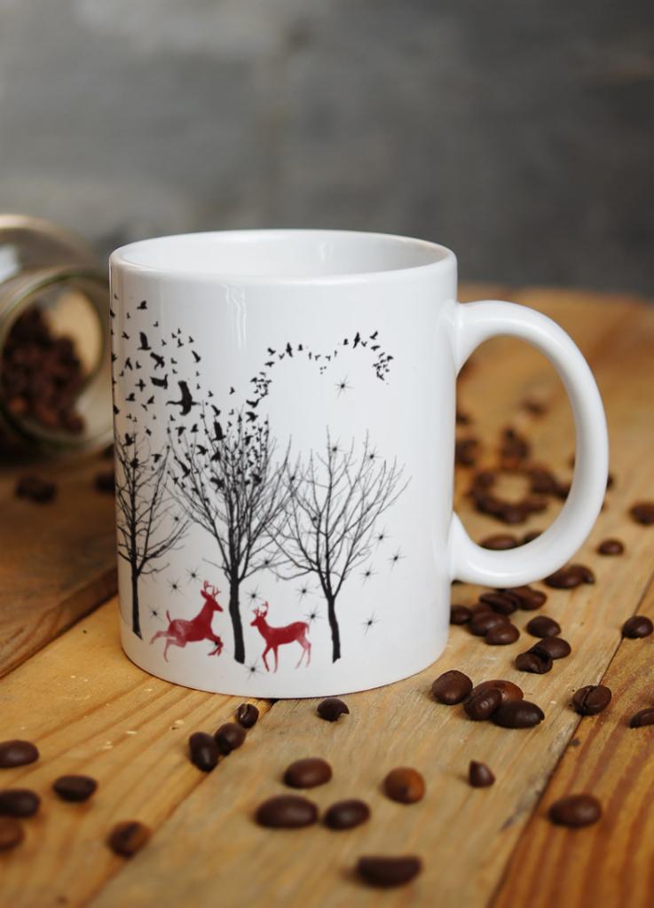 Biały kubek z nadrukiem zimowego krajobrazu: sylwetki drzew, stado ptaków i dwa czerwone jelenie, otoczony rozsypanymi ziarnami kawy na drewnianym stole.