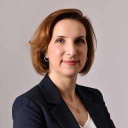 Małgorzata Sobotka Agent Ubezpieczeniowy - Portret kobiety w granatowej marynarce, z delikatnym uśmiechem i subtelną biżuterią, na jasnoszarym tle. Profesjonalny wizerunek.