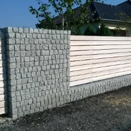 Nowoczesne ogrodzenie z jasnego drewna i kostki granitowej na tle domu jednorodzinnego w słoneczny dzień. Widok na fragment ogrodzenia.