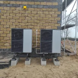 Dwie szare pompy ciepła Viessmann zamontowane na betonowych podstawach przed budynkiem z cegły klinkierowej w trakcie prac wykończeniowych, widoczne rusztowania i piasek.