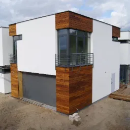 Nowoczesny dom z białą elewacją i drewnianymi panelami, balkon z metalową balustradą, garaż z szarą bramą. Widoczne elementy wykończenia i otoczenie domu.