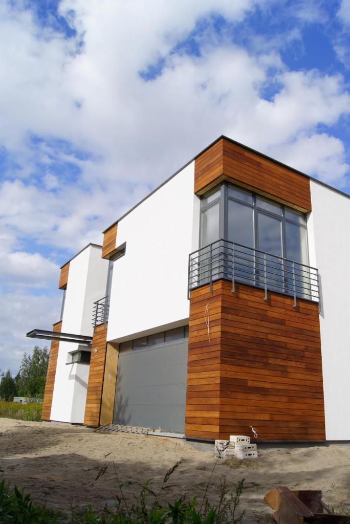 Nowoczesny dom z białą elewacją i drewnianymi panelami, balkon z metalową barierką. Widok z dołu na budynek w słoneczny dzień.