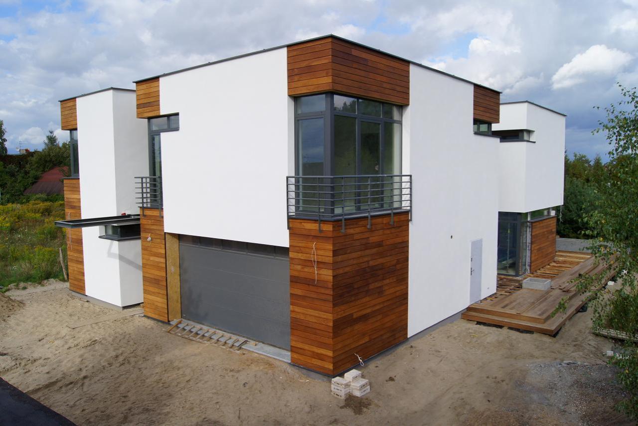 Nowoczesny dom z białą elewacją i drewnianymi panelami, balkon z metalową balustradą, garaż z szarą bramą. Widoczne elementy wykończenia i otoczenie domu.