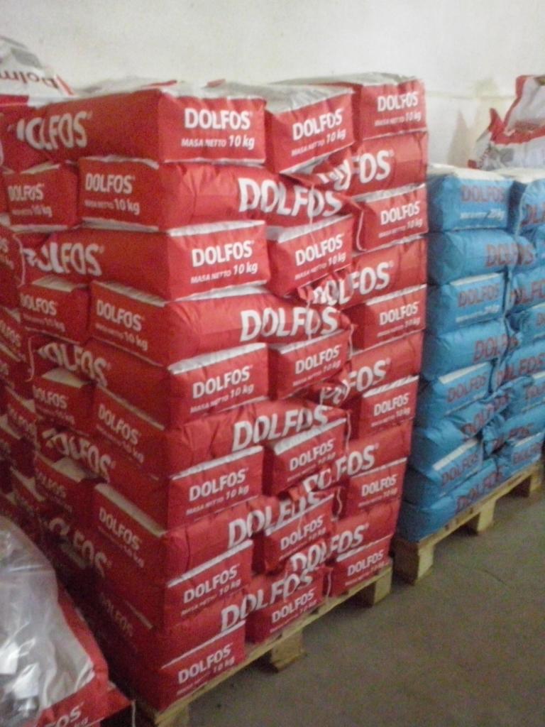 Stos czerwonych worków z napisem DOLFOS (masa netto 10 kg) na paletach, obok niebieskie worki. Magazyn z zapasami.