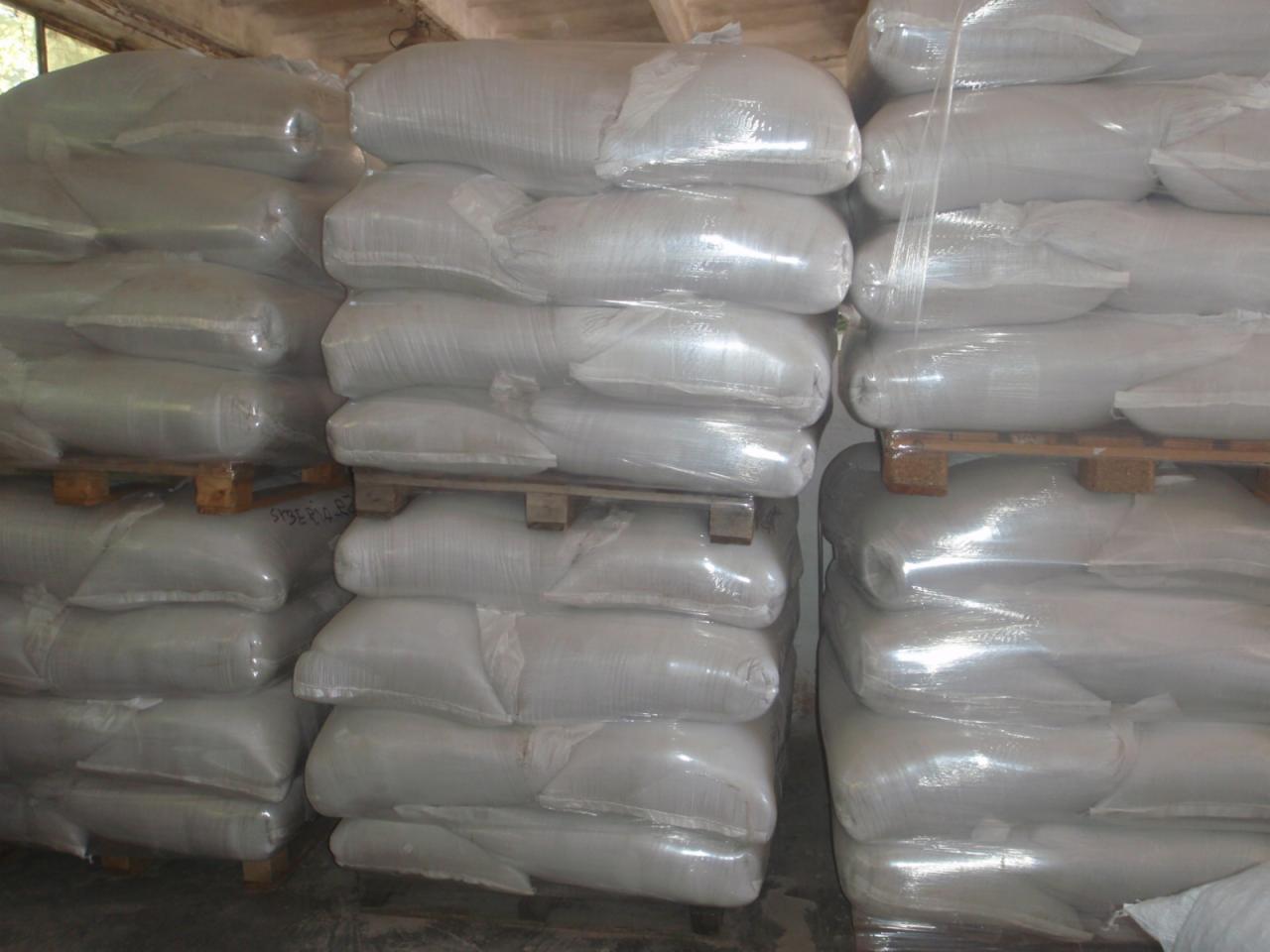 Stosy białych worków na paletach, zabezpieczonych folią, w magazynie. Widoczne napisy na jednym z worków. Oświetlenie sufitowe.