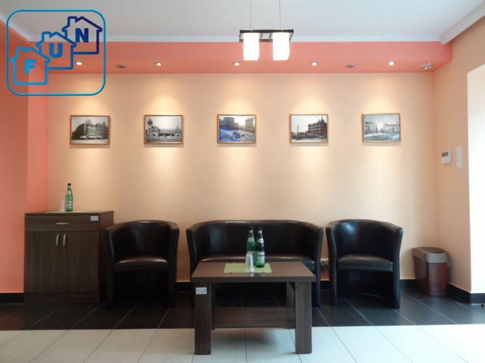 Poczekalnia biura z ciemnymi fotelami, sofą i stolikiem kawowym, na ścianie wiszą zdjęcia budynków, logo firmy w lewym górnym rogu.