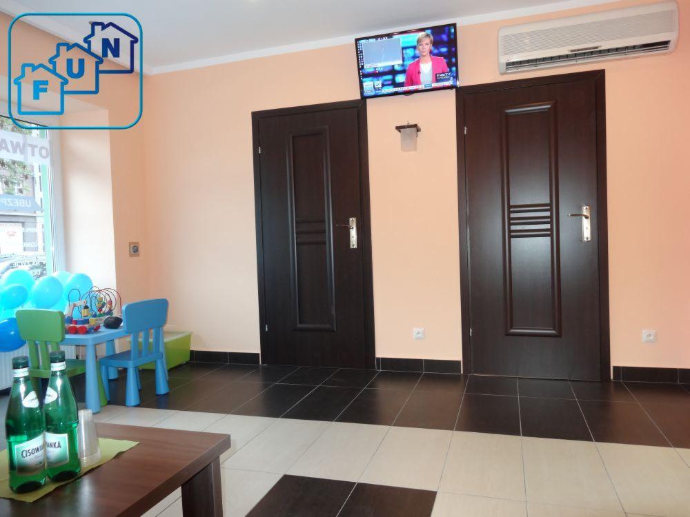 Wnętrze biura z dwoma ciemnymi drzwiami, telewizorem na ścianie, stolikiem z zabawkami dla dzieci i butelkami wody mineralnej na pierwszym planie, logo firmy w lewym górnym rogu.